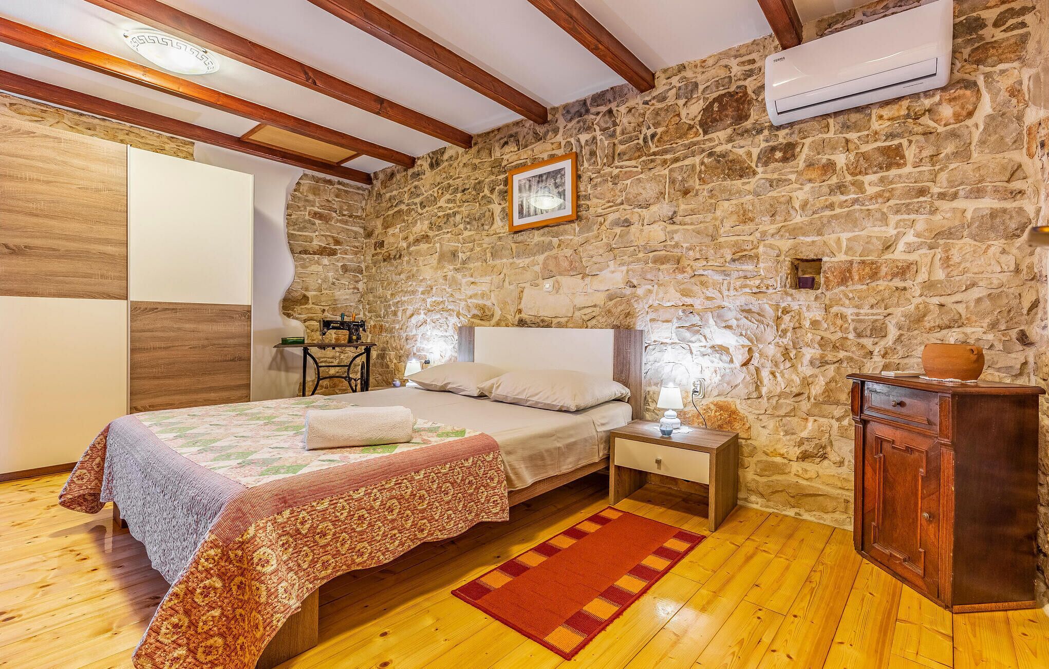 Une chambre avec un mur en pierre, des poutres apparentes, un lit recouvert d’une couvre-lit à motifs, une table de chevet, une commode et un appareil de climatisation.