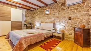 Camera da letto con parete in pietra, travi in legno, un letto con copripiumino decorato, un comodino, una cassettiera e un condizionatore d'aria.