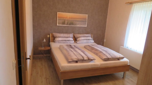 A bedroom with a wooden bed, a bedside table with a lamp, a painting on the wall, and a window with curtains.