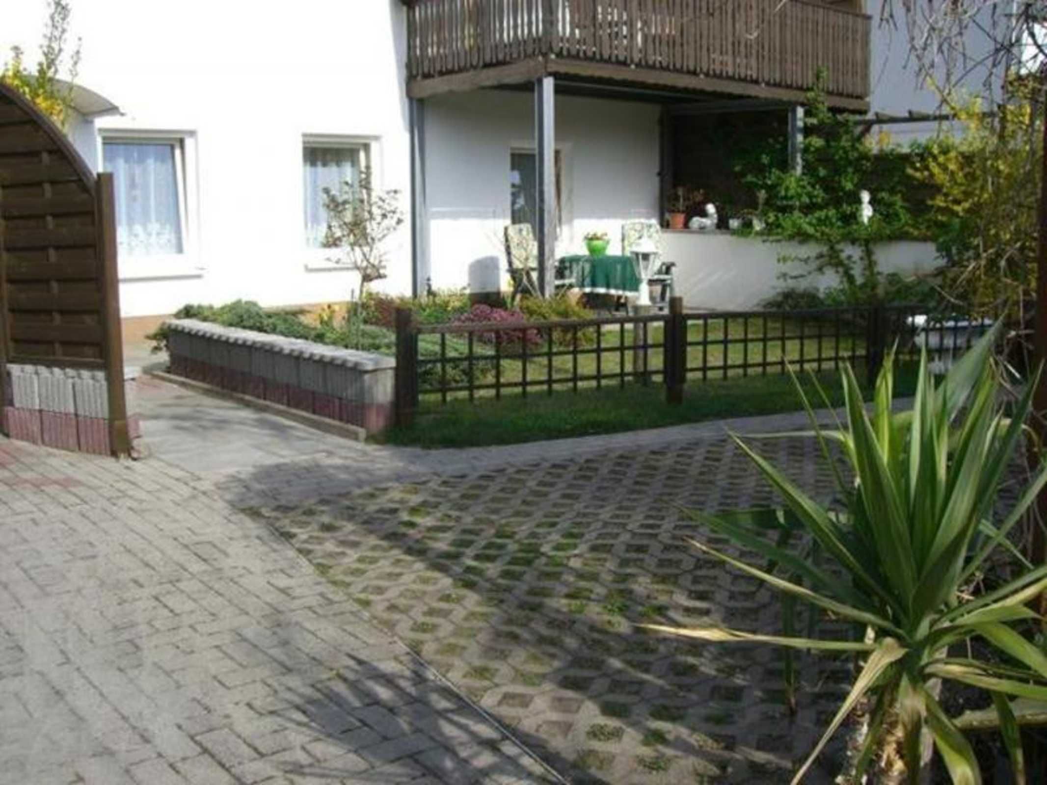 Edifici residencial amb un balcó, una entrada pavimentada i una zona de jardí amb plantes i una tanca.