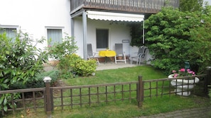A covered patio with a yellow table and chairs, surrounded by greenery and a wooden fence.