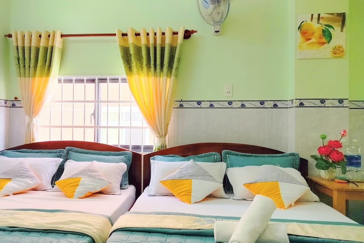 Une chambre d’hôtel avec deux lits, chacun agrémenté de plusieurs oreillers et recouvert d’une courtepointe matelassée. On y trouve une fenêtre avec des rideaux, un ventilateur fixé au mur et une petite table sur laquelle est posé un vase de fleurs.