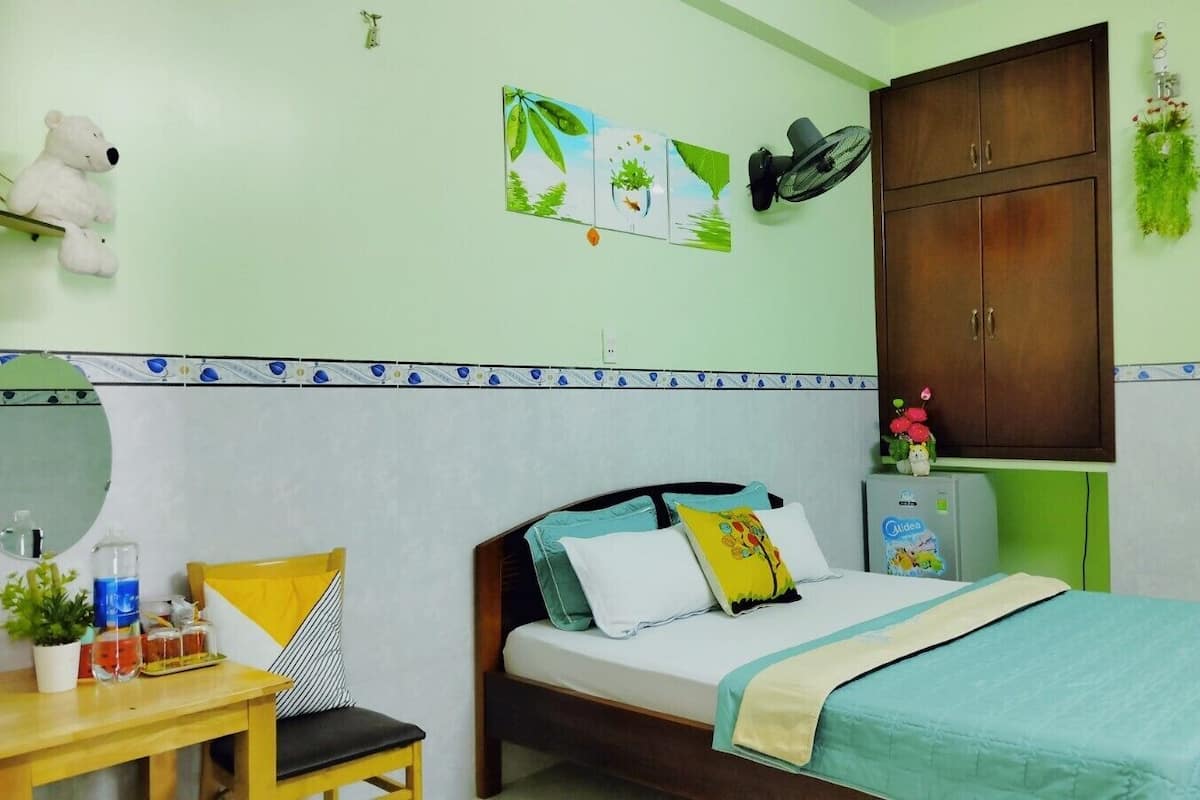 Une chambre avec un lit, un bureau avec une chaise, un miroir, un petit réfrigérateur et un ventilateur fixé au mur.