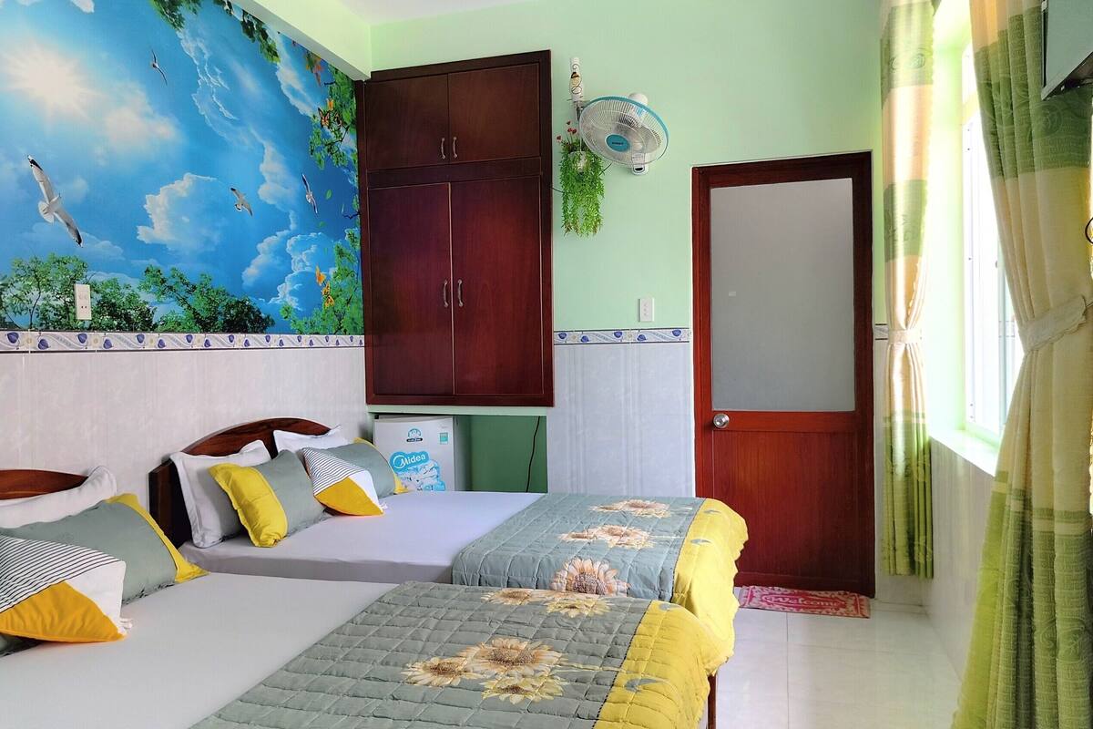 Une chambre avec une fresque représentant des oiseaux et des arbres, une armoire, un lit avec de nombreux oreillers et une table de chevet avec un ventilateur.