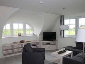 A modern living room with a flat-screen TV, a sofa, a coffee table, and a lamp. There are books and decorative items on shelves, and a window with a view of greenery. - Vacation home Hiddensee on the Baltic Sea - Apartment III (Meeresrauschen) (Insel Hiddensee)