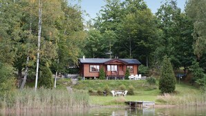 Une cabane avec une terrasse et un petit quai au bord d’un lac, entourée d’arbres.