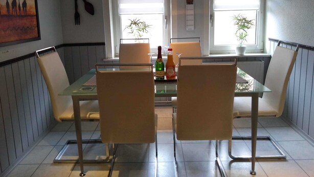 Zona pranzo con tavolo in vetro e quattro sedie, due finestre con piante e un quadro appeso al muro.