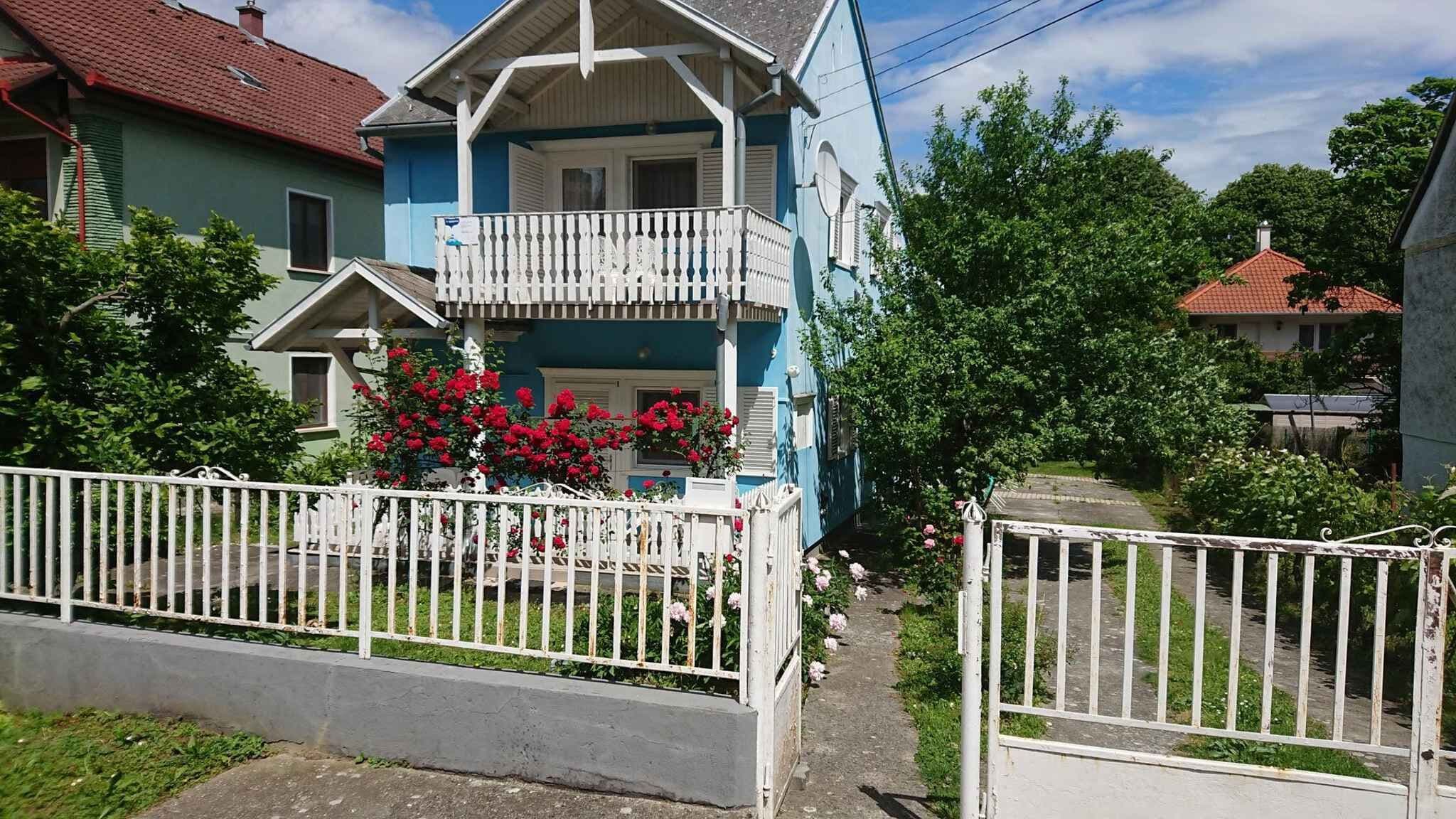 A two-story house with a balcony, surrounded by a white fence and lush greenery.