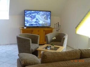A living room with a television on a wooden cabinet, two armchairs, a small coffee table, and a staircase with a yellow light.