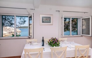 Een eetruimte met een tafel gedekt voor vier personen, twee ramen met gordijnen, een ingelijst schilderij aan de muur, en uitzicht op zee en boten.