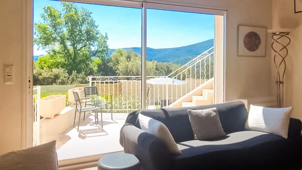 Ein modernes Wohnzimmer mit einem dunklen Sofa, weißen Kissen und Blick auf einen Balkon mit Geländer und einem Stuhl.