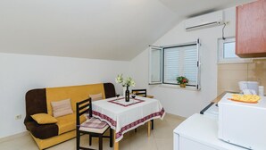 A small, cozy room with a yellow sofa, a dining table with a white tablecloth, a window with shutters, a white refrigerator, and a wall-mounted air conditioner.