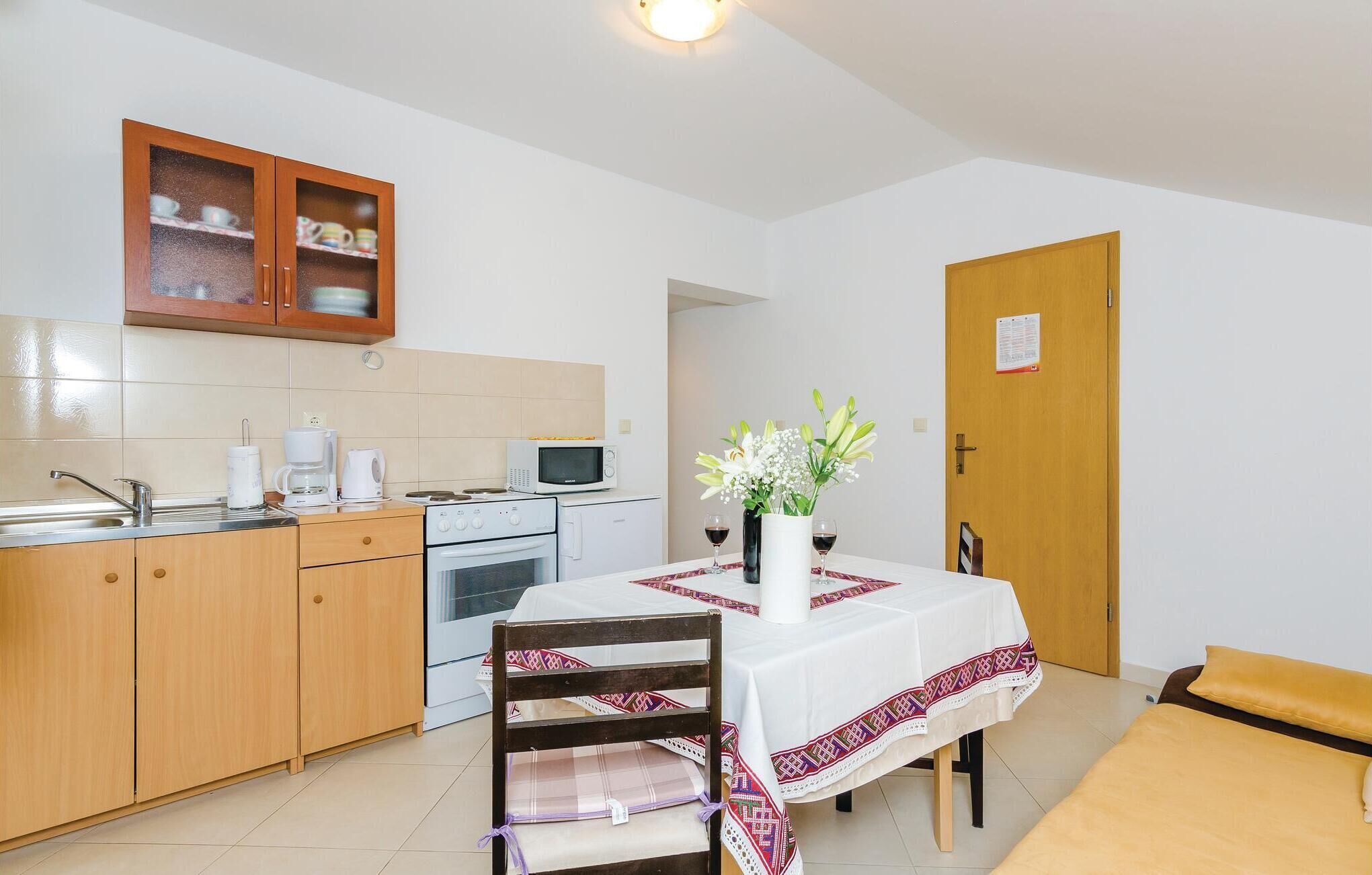 Una cocina compacta con microondas, estufa y armarios de madera. Una mesa de comedor con mantel blanco y un jarrón de flores dispuesta para dos comensales.
