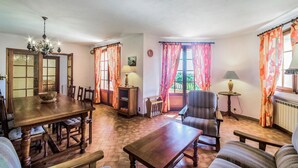A cozy living room with a wooden dining table, a sofa, and a chair. There is a chandelier, a lamp, and curtains on the windows. - Domaine de la Bastide d'Agay (Agay)