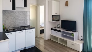 Une cuisine compacte équipée d’une cuisinière à gaz, d’un évier et de meubles blancs. Un téléviseur est fixé sur un meuble blanc, avec une étagère en dessous.
