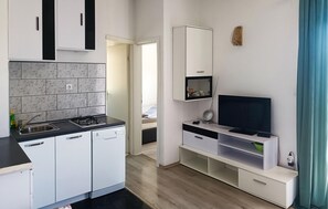 A compact kitchen with a gas stove, sink, and white cabinetry. A TV is mounted on a white stand with a shelf below. - 2 bedroom gorgeous apartment in Split (Split)