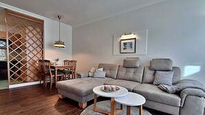 Un salon moderne avec un canapé d’angle gris, une table à manger en bois et des chaises. Une table basse ronde, avec un plateau blanc, est placée au centre.