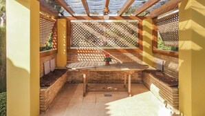 Een overdekt terras met een houten tafel en bankjes, omgeven door bakstenen muren en lattenwerkpanelen.