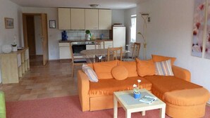 Un salon avec un canapé orange, une petite table sur laquelle est posée une plante, et une cuisine à l’arrière-plan.