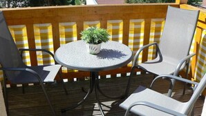 Une terrasse en bois avec une table ronde et deux chaises, une plante en pot posée sur la table, et une clôture jaune et blanche à rayures en arrière-plan.