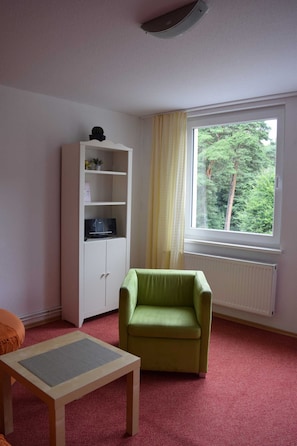 Une pièce avec un fauteuil vert, une armoire blanche, une petite table et une fenêtre donnant sur des arbres.