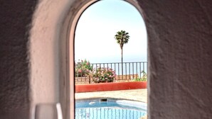 A view from a room with a tiled floor and a glass table, overlooking a swimming pool with a palm tree and a fence in the background.