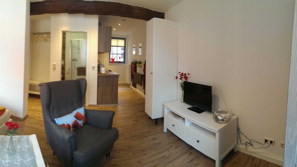 Una habitación de hotel moderna con una cama grande, dos sillones, una mesita, un televisor y una kitchenette al fondo.
