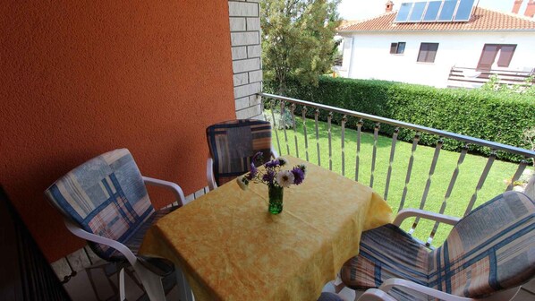 Un pequeño balcón al aire libre con una mesa y sillas, un florero con flores y vistas a un jardín y una casa al fondo.