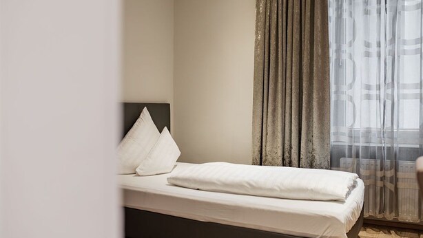 Una cama bien hecha con sábanas blancas, cabecero oscuro y una ventana con cortinas translúcidas.