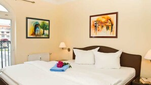 A hotel room with a bed, two paintings on the walls, a nightstand with a lamp, and a window with a view of buildings. - (1) 3 room apartment - sea views - apartment block "Atlantic" (Kühlungsborn)