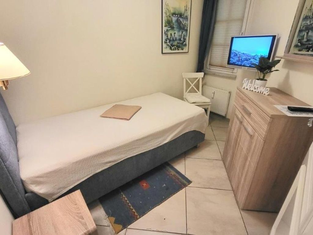 A small bedroom with a single bed, a desk with a 'Welcome' sign, a television, and a window with blinds.