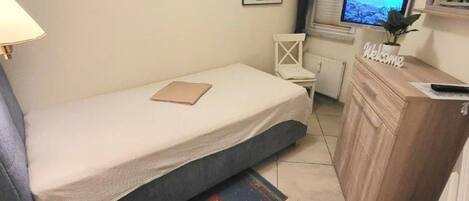 A small bedroom with a single bed, a desk with a 'Welcome' sign, a television, and a window with blinds.