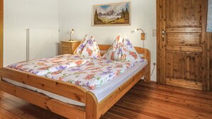 Een houten bed met bloemenbedekking, een nachtkastje met een lamp, een schilderij van een berglandschap, en een houten deur.