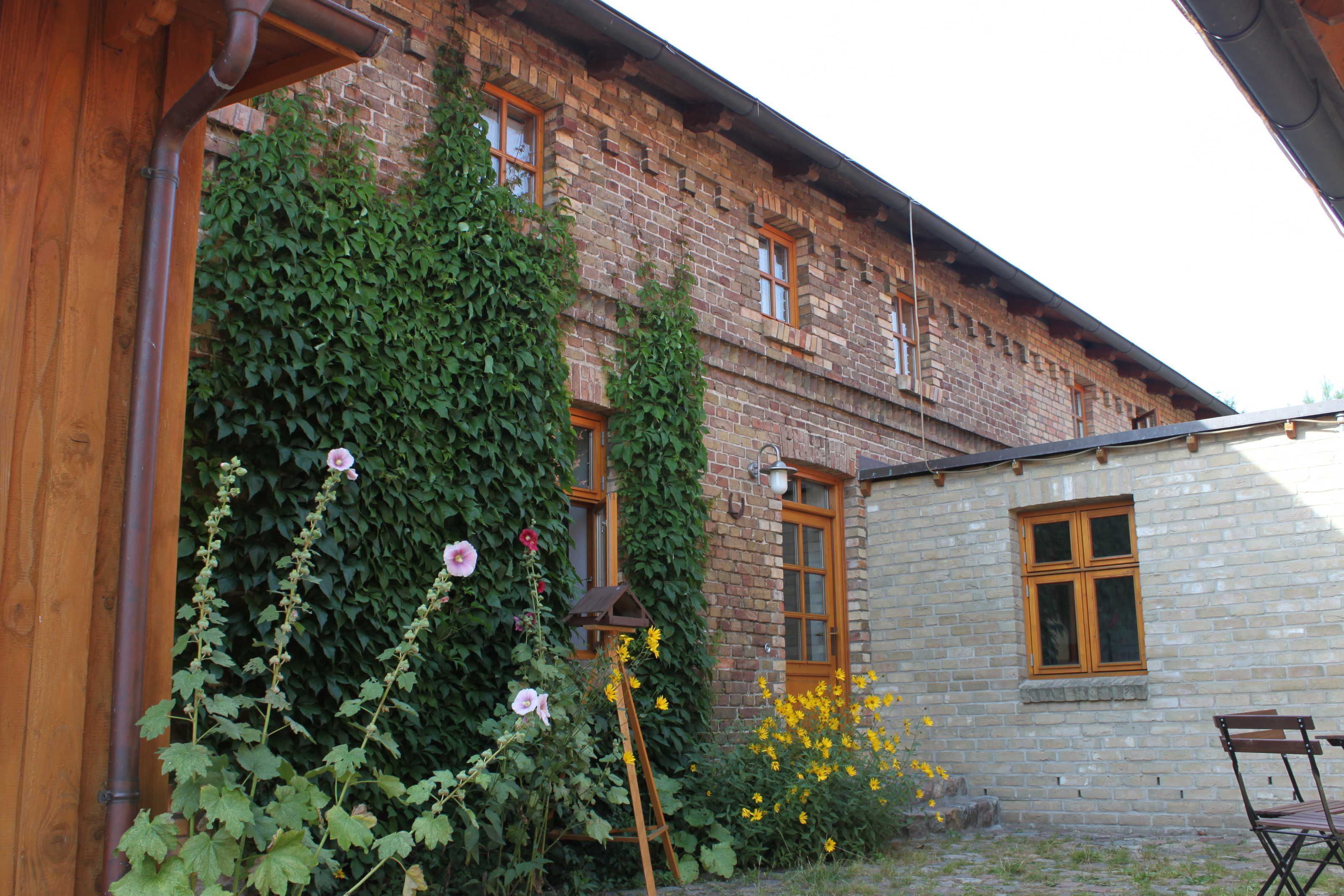 A brick building with ivy-covered walls, a wooden structure on the left, and a small table and chair on the right.