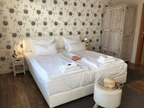 A double bed with white linens, a nightstand with a lamp, and a wardrobe.
