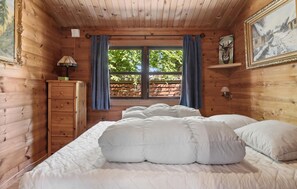 Ein Schlafzimmer einer Holzhütte mit einem Bett, einer Kommode, einem Fenster mit Vorhängen und einem Bild an der Wand.