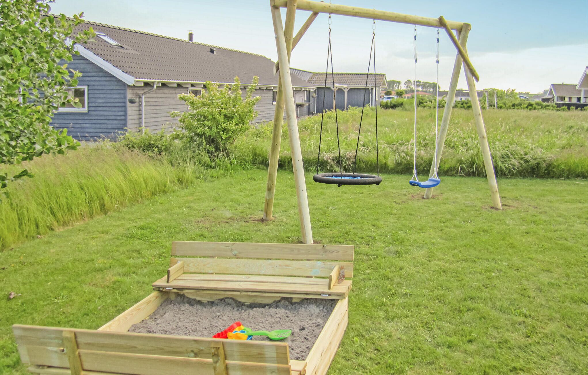 A wooden swing set with two swings, a sandbox, and a grassy yard with a house in the background.