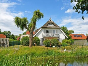 A white house with a balcony, surrounded by greenery and a pond.