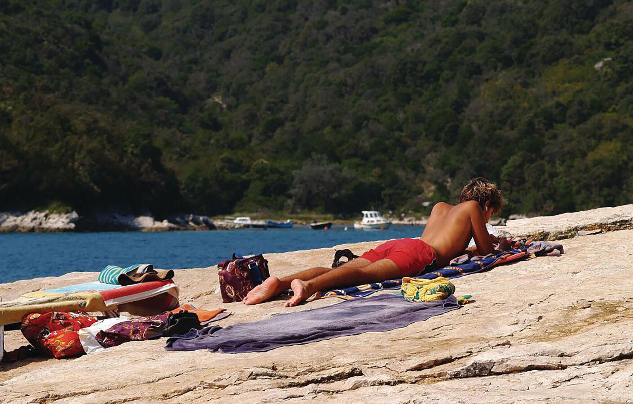 A person sunbathing on a rocky beach with a bag and clothing items nearby.