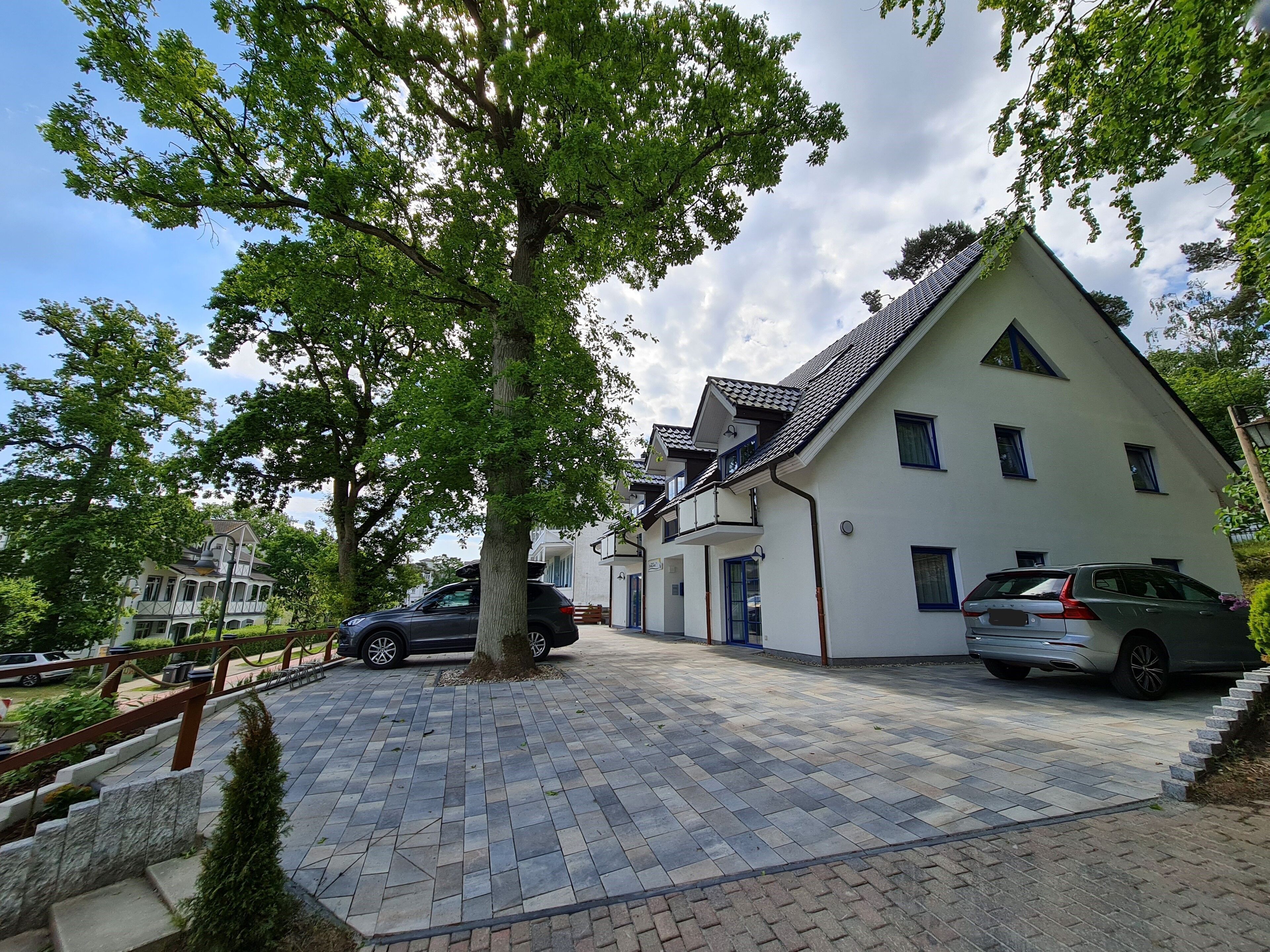 Une maison à deux étages avec une allée carrossable dallée et un grand arbre devant.