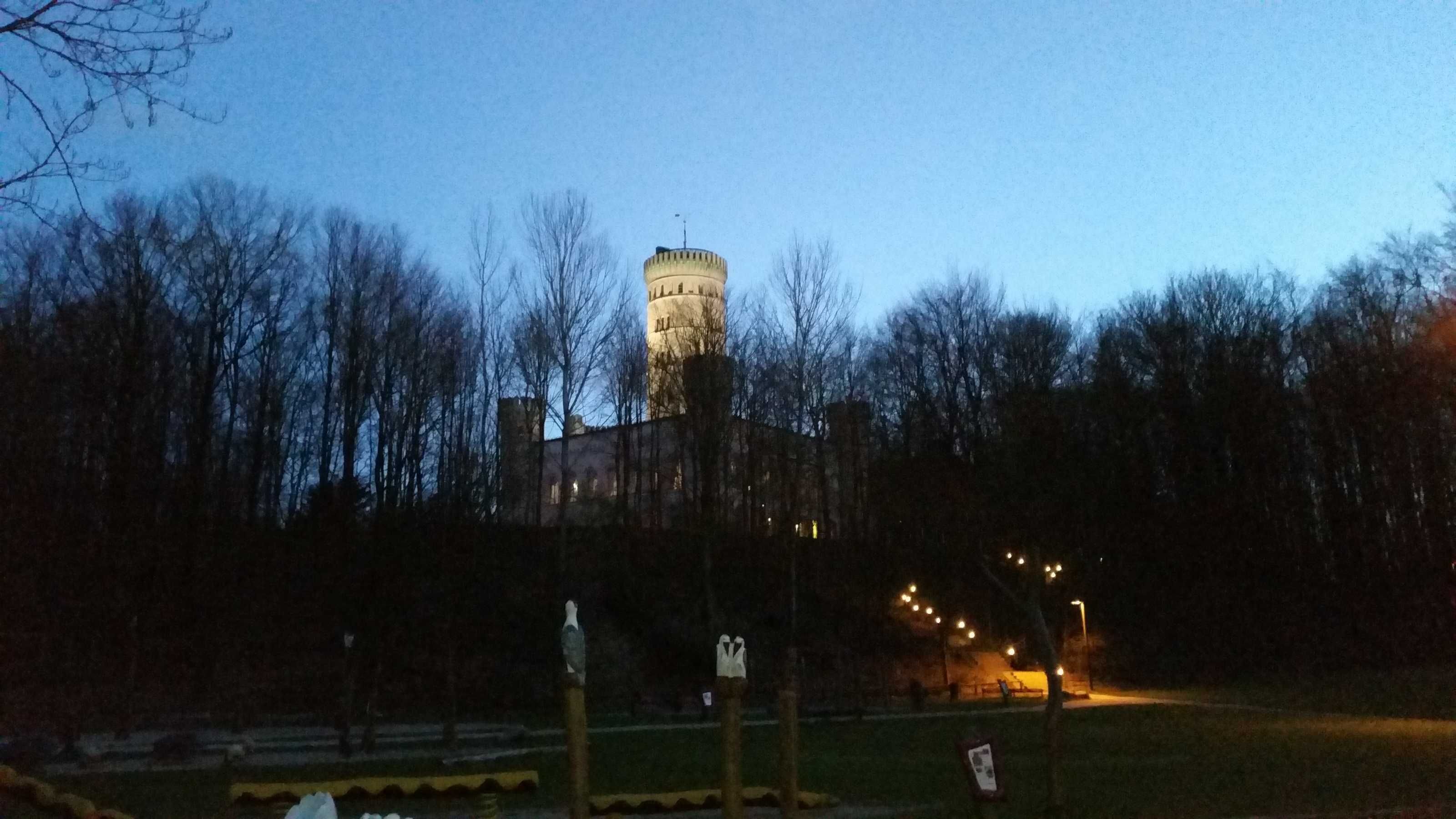 A tower with a clock, surrounded by trees and a park area with benches and lights.