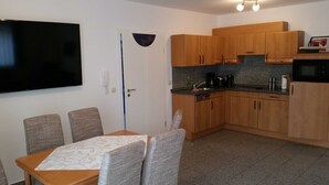 Una cocina compacta con armarios de madera, un microondas y una cafetera. Hay una mesa de comedor con cuatro sillas y un televisor montado en la pared.