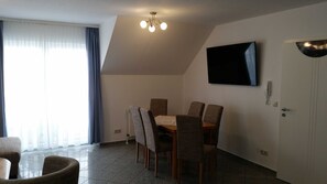 Un coin repas avec une table et des chaises, un téléviseur à écran plat fixé au mur, et une porte équipée d’un téléphone et d’un interphone.
