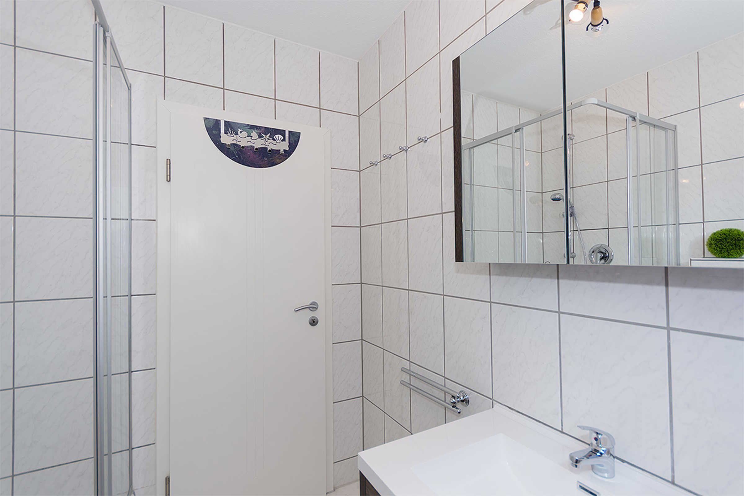 Une salle de bain moderne avec des carreaux blancs, une douche en verre, un lavabo avec un miroir et une petite plante verte posée sur le comptoir.