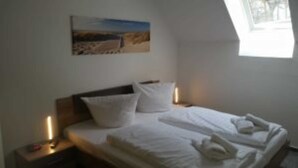 Una cama bien hecha con sábanas y almohadas blancas, una mesita de noche con una lámpara, y un cuadro en la pared.