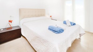 A neatly made bed with white bedding and blue towels, a bedside table with a lamp, and a window with curtains. - 2 bedroom gorgeous apartment in Roldán (Roldán)