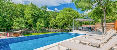 A swimming pool with lounge chairs, a table, and a covered seating area surrounded by trees.