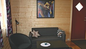 A room with a wooden wall, a black sofa, a black armchair, a small table with a doily, a lamp, and a framed abstract painting. - 2 bedroom stunning apartment in Sandstad (Sandstad)
