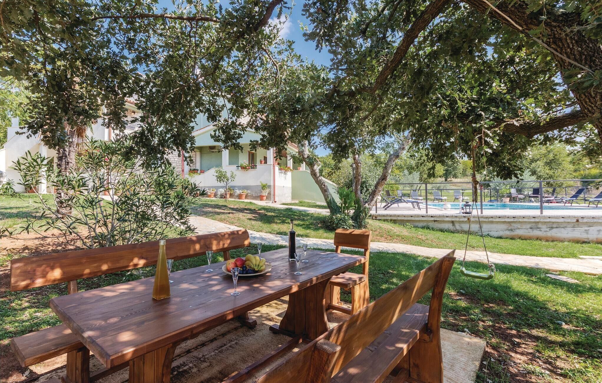 Une table en bois installée à l’extérieur, avec des bancs, entourée d’une végétation luxuriante et, au fond, une zone avec une piscine.