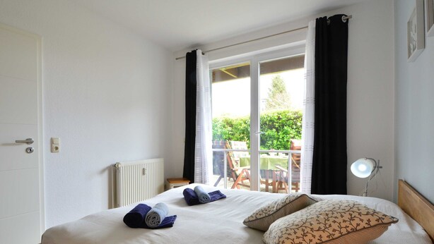 A bedroom with a bed, a radiator, a window with curtains, and a view of an outdoor area with chairs and a table. - Wohnpark An der Muehle Whg Cuba1-1 .. - Cuba 1. 1 (Kühlungsborn)